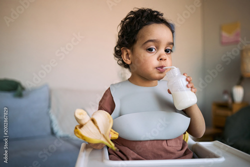Fotografi Portrait of toddler drinking milk from bottle while holding banana in high chair