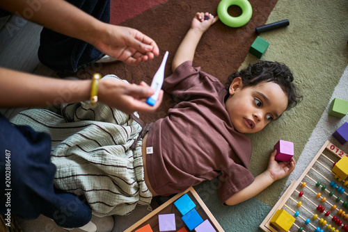 Tableau sur toile Overhead shot of toddler lying on play mat while parent checking thermometer, su