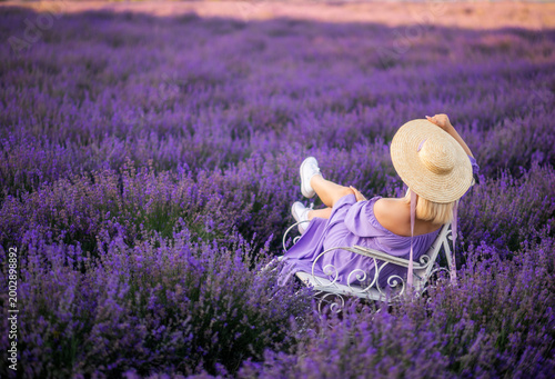 woman sits in a field of lavender on a rocking chair. A braided hat and purple flowers all around . View from the back