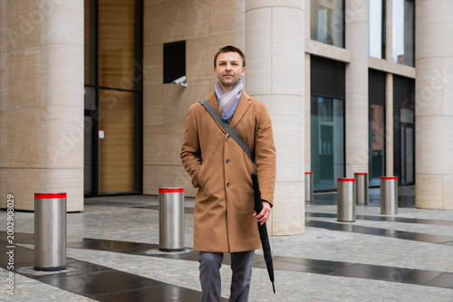 Confident man in brown coat standing near modern business center. Calm lawyer or finance professional holding umbrella. Serious city inhabitant outdoors. Successful career and lifestyle concept.