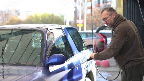 Slow motion 4K footage of a man washes a blue car at a self-service car wash. The car wash's high-pressure jet sprays water and rinses away the cleaning foam.