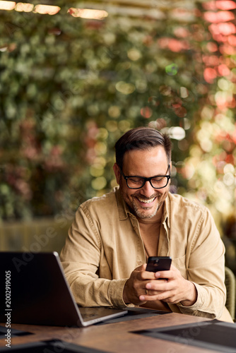 Smiling Man Working Remotely on Laptop and Phone at Outdoor Cafe, Casual Business Lifestyle