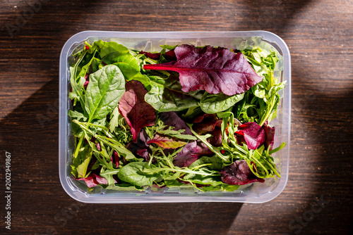 Fresh mixed salad leaves in plastic container on wooden background. Organic salad mix with spinach arugula and beet leaves in transparent plastic box on dark wooden table with hard shadows, top view