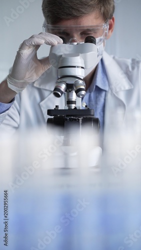 Obraz na płótnie Man scientist wearing lab coat, white gloves, medical mask and protective glasses using microscope doing research in modern laboratory, portrait view