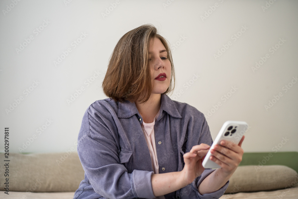 Naklejka premium Plus size woman using smartphone on sofa at home, mobile lifestyle, messaging, online communication, social media, digital routine, modern living, body positivity.