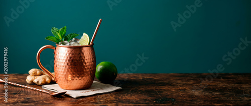 Moscow mule cocktail served in a hammered copper mug adorned with fresh lime and mint garnishes rests on a rustic wooden table alongside ginger and a spoon illuminated by studio lighting