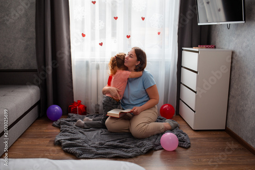 Wide shot of little Caucasian girl hugs her mother, sits on a carpet in room and reads book. Woman and her daughter spend happy time. Mother's Day Concept