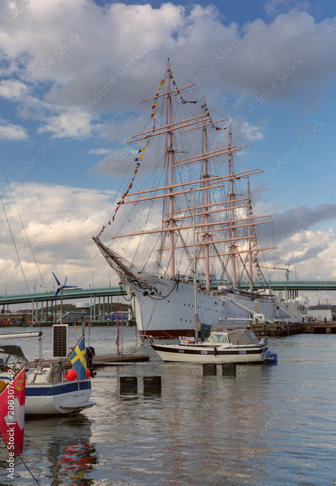 Fototapeta premium Tall ship moored in Gothenburg harbor with waterfront skyline, marina boats and dramatic evening clouds in Sweden.