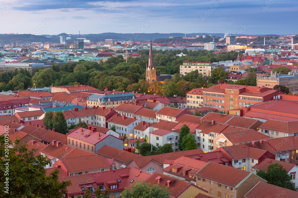 Obraz premium View of illuminated church spire rising above Gothenburg rooftops and green parkland at dusk in Sweden.