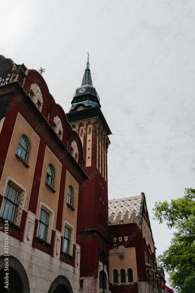 Obraz premium The clock tower and facade of Subotica City Hall rise above the street in Subotica, Serbia. Historic architecture, civic landmark, urban tourism, and European heritage.