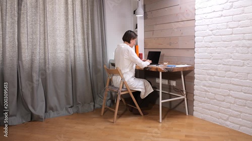 Female professional wearing a lab coat and sitting at a wooden desk, focused on typing on a laptop, representing remote work and healthcare or scientific research