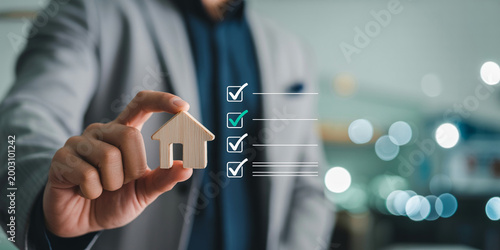 Professional real estate agent holding small wooden house model with digital property checklist.
