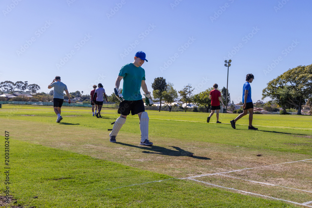 Obraz premium Male teammates walking across marked cricket pitch in park wearing batting pads and gloves
