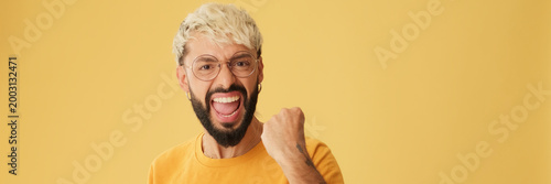 Guy with glasses, dressed in yellow T-shirt, getting good news, celebrates victory, isolated on yellow background in studio. Panoramic. Panoramic