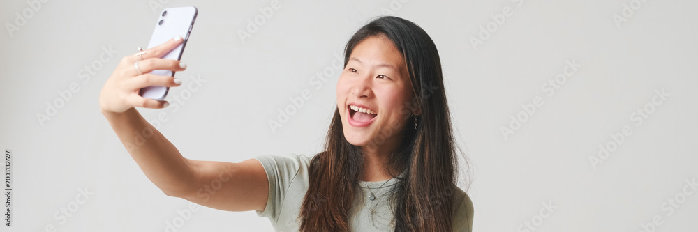 Fototapeta premium Smiling girl in gray top takes selfie on mobile phone, makes video call, isolated on white background, studio shot. Panoramic