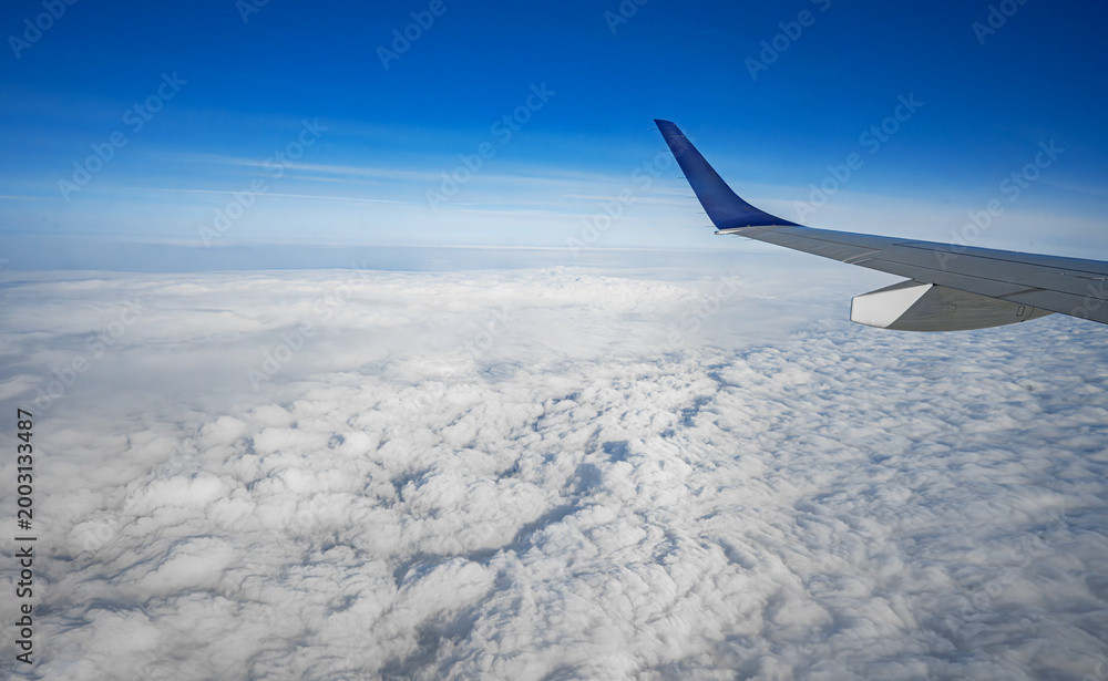 Obraz premium An aerial view from an airplane window shows a silver wing soaring above a thick blanket of white clouds under a clear blue sky.