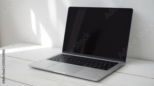 A modern silver laptop with a dark screen, placed on a white wooden table against a minimalist white wall with soft sunlight creating subtle shadows.