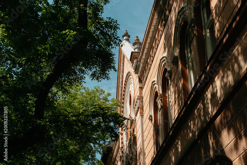 Historic townhouse facade framed by leafy trees in Subotica, Serbia. Ornate residential architecture in warm summer light.