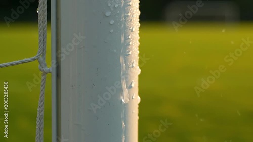 Macro shot of water droplets dripping down a white soccer goal post with a blurred green grass field background in golden sunlight.