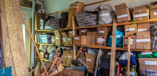 Schilderij op canvas A cluttered, messy wooden shelf in a storage area is filled with disorganized cardboard boxes, plastic bags, plaid storage bags, and various loose items, reflecting a busy or untidy environment