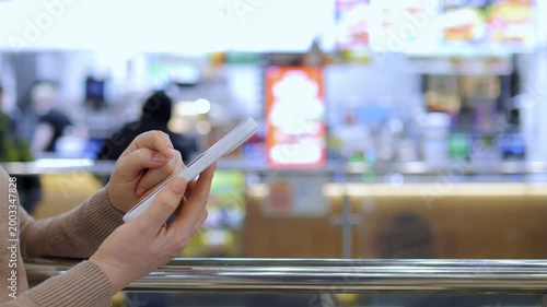 Person holds phone, typing at counter. Background shows blurred food stall. Focus on hands and device. Scene indoors, likely cafeteria or market. No animals or plants visible