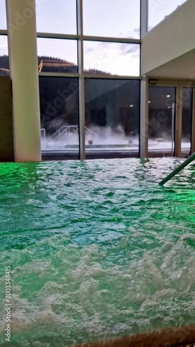 Bubbling turquoise water in a jacuzzi with colorful underwater lights, overlooking a foggy mountain landscape through large windows. Concept of relaxation and natural beauty intertwined.