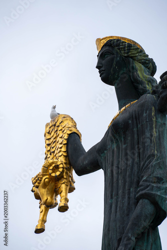 Low angle view of a dark bronze statue with golden details holding a ram, against a pale sky, with a seagull perched on the sculpture.