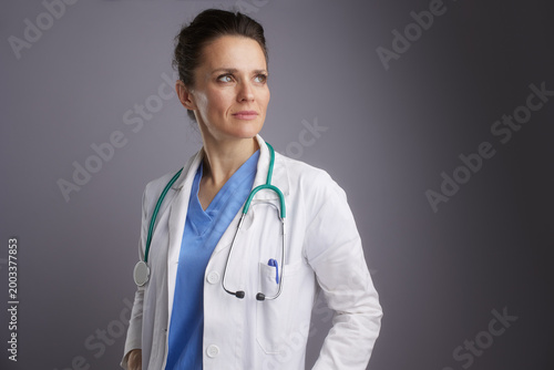 Portrait of a focused female medical professional wearing blue scrubs and a white lab coat, conveying expertise and commitment to patient care.