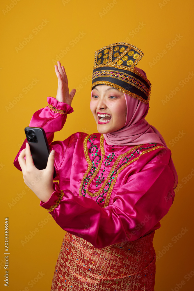 Naklejka premium Excited Young Indonesian Asian Woman in Hijab and Traditional Costume Taking a Selfie on Yellow Background