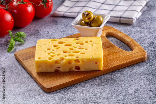 Block of Swiss cheese on a wooden cutting board with grilled olives and fresh tomatoes