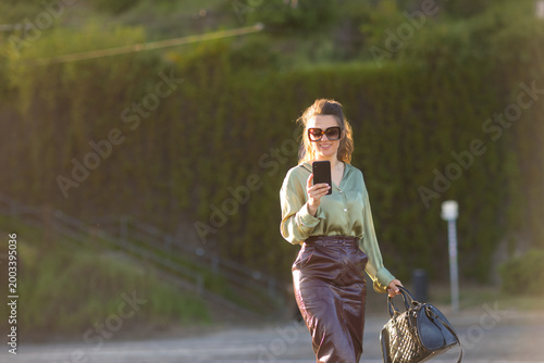 A cheerful, stylish woman in a green blouse and burgundy leather skirt uses her smartphone outdoors during the warm glow of golden hour in the city.