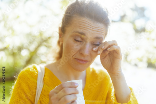 A middle-aged woman expresses discomfort while rubbing her eyes in a bright spring setting. Her struggle with tree pollen allergies is highlighted against a background of green foliage.