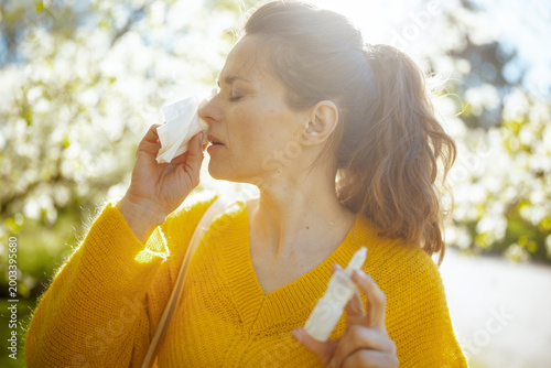 Backlit by warm spring sunlight, a woman in a yellow sweater manages hay fever symptoms with a tissue and nasal spray amidst flowering trees.