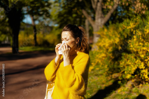 A woman in a yellow sweater uses a tissue while sneezing in a sunlit park, capturing the discomfort of seasonal pollen allergies during the spring bloom.