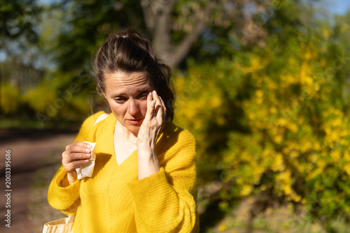 A woman in a yellow sweater rubs her irritated eyes while holding a tissue, depicting common hay fever and pollen allergy symptoms in a spring park.