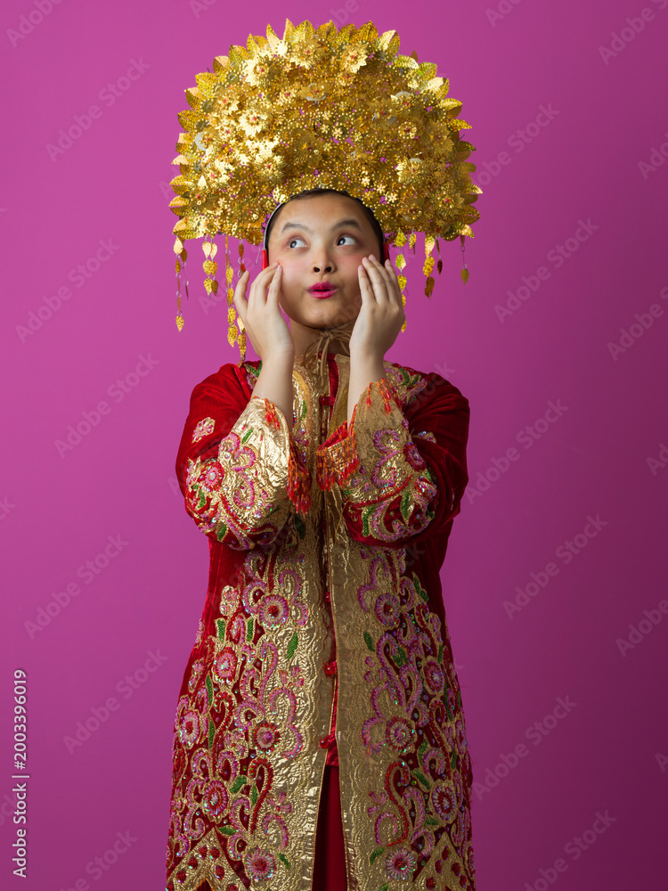 Fototapeta premium Young Indonesian Asian Woman in Ornate Gold Headdress and Embroidered Red Traditional Costume on Pink Background