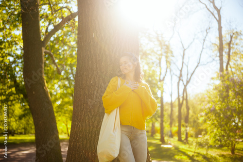A woman in a yellow sweater leans against a tree, hands over her chest, embracing serenity and connection with nature in a sun-drenched forest park.