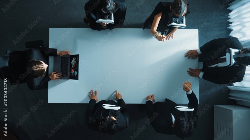 Fototapeta premium Top down aerial view of diverse data analysts wearing VR headset and stand around table while using hand gestures to manipulate the data streamed live from executive manager's laptop. Directorate.