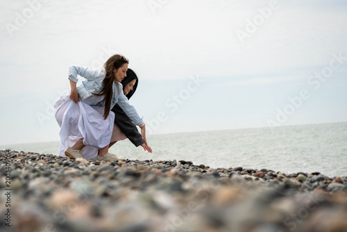 Fotografija Two young women crouching on a pebble beach by the sea, touching stones and enjoying a quiet coastal moment under an overcast sky