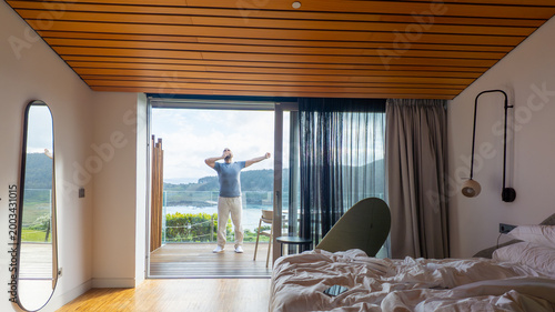 Luxury Modern Bedroom with Unmade Bed and Man Stretching on Balcony Overlooking Coastal Landscape
