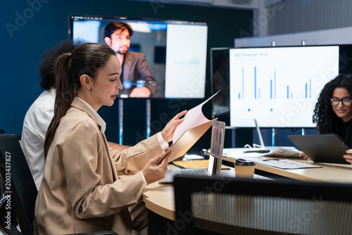 Young businesswoman reviewing documents during a hybrid meeting about AGI financial trends. Professional female executive presenting data on digital screens with a diverse team in a modern office