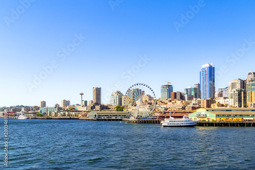 The Seattle Great Wheel and the bustling waterfront piers take center stage against a backdrop of downtown skyscrapers and the distant Space Needle, all bordering the blue waters of Elliott Bay