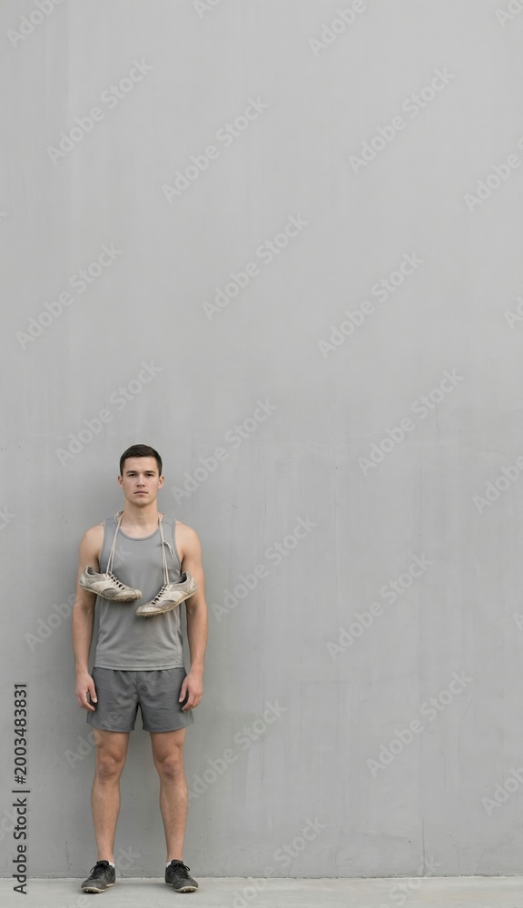 Fototapeta premium Male athlete in sportswear with shoes around neck standing against gray wall