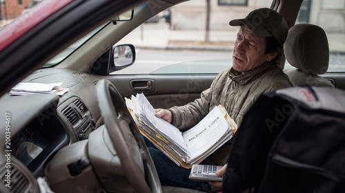 Man sitting inside car reviewing mileage logs with calculator representing tax deductions expense tracking delivery work self employment audit preparation or financial recordkeeping challenge concept