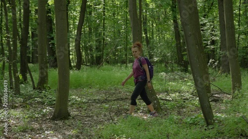 A woman with curly red hair carrying a purple backpack walks away along a dirt trail through dense green spring woodland