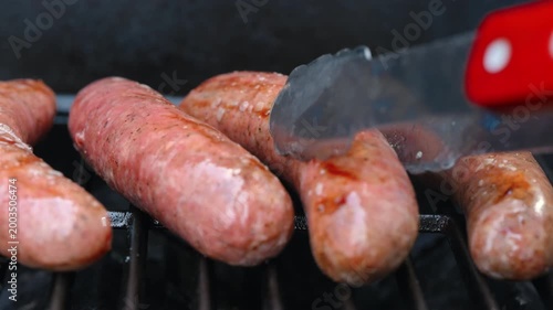 View of raw sausages turning over while grilling on barbecue with open flame. Close up of juicy meat cooking on grill, outdoor food and summer barbecue
