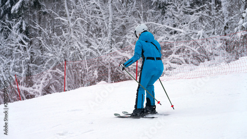 Fényképezés An inexperienced skier in a blue jumpsuit skis down a snowy slope against a picturesque backdrop of snow-covered trees