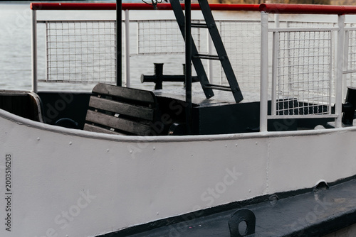 A small ferry boat docked by the waterfront on a cloudy day, with visible details like a lifebuoy and rope along the deck.