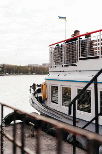 A small ferry boat docked by the waterfront on a cloudy day, with visible details like a lifebuoy and rope along the deck.