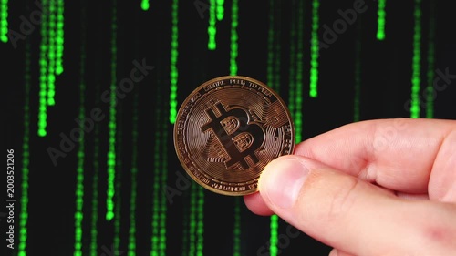 Une pièce Bitcoin tenue par une main, devant un fond de code vert, symbolisant la technologie blockchain, la cybersécurité et l’univers des cryptomonnaies.